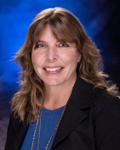 Woman with long wavy brown hair and curtain bangs wears a navy blue shirt under a black blazer